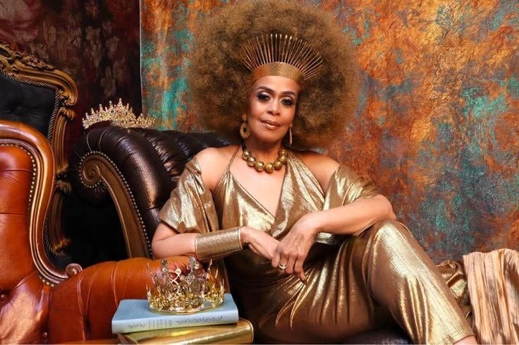 Marsha — gold metallic jumpsuit, seated on leather chair, copper teal backdrop, crown